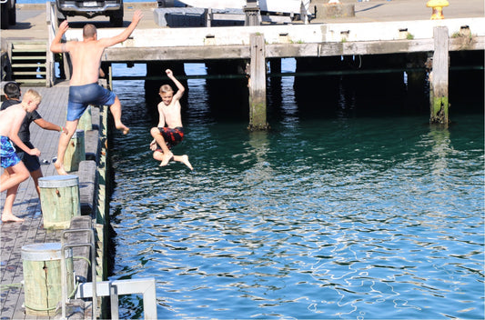 "A group of boys jumping off a wooden dock into the clear blue water, with one boy in mid-air and others preparing to jump, enjoying a sunny day at the waterfront.