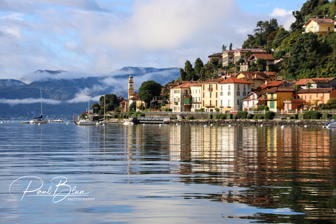 Scenic view of a lakeside village on Lake Maggiore, Italy, with colourful buildings, a church tower, and boats moored along the calm, reflective water, set against a backdrop of misty mountains.