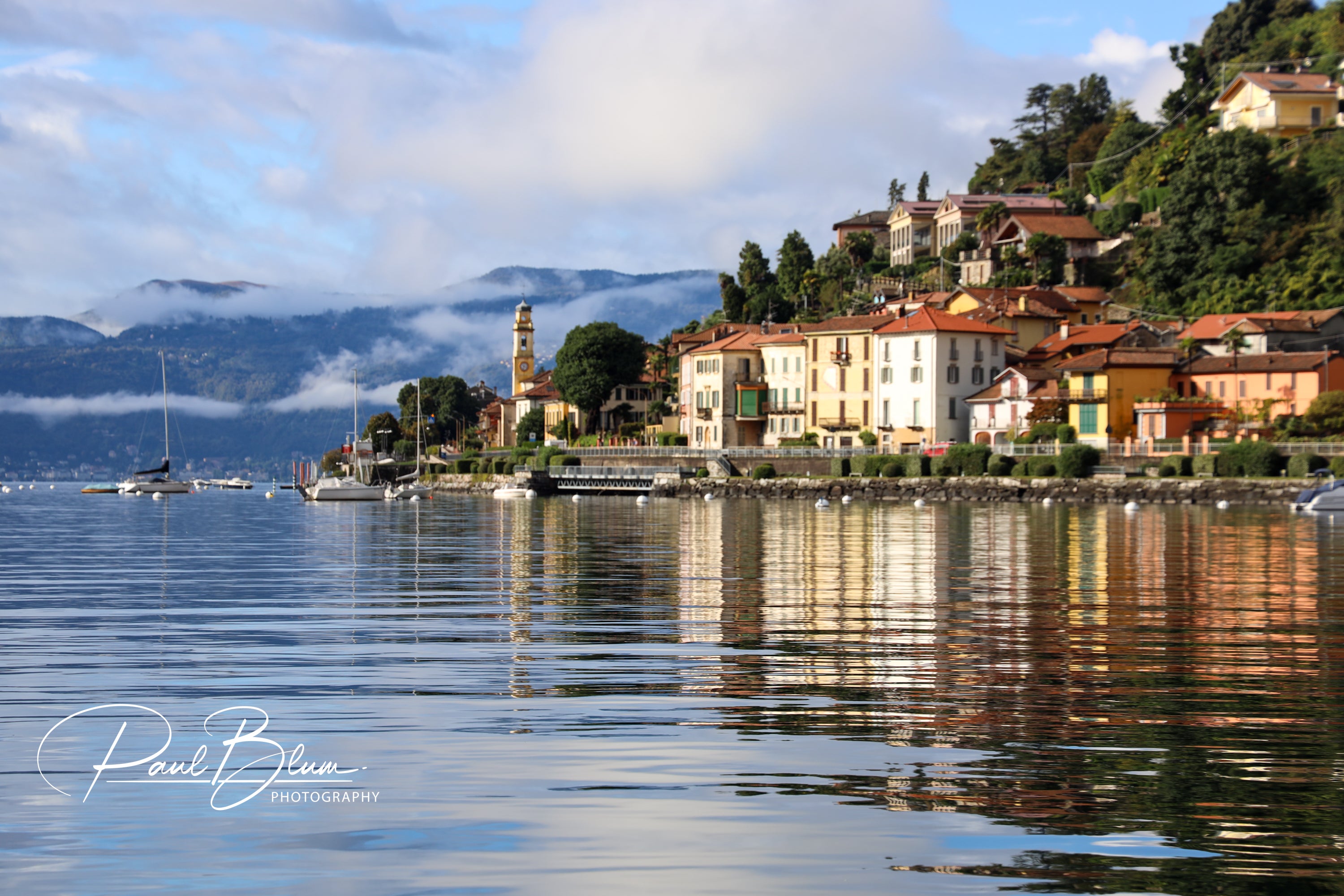 Scenic view of a lakeside village on Lake Maggiore, Italy, with colourful buildings, a church tower, and boats moored along the calm, reflective water, set against a backdrop of misty mountains.