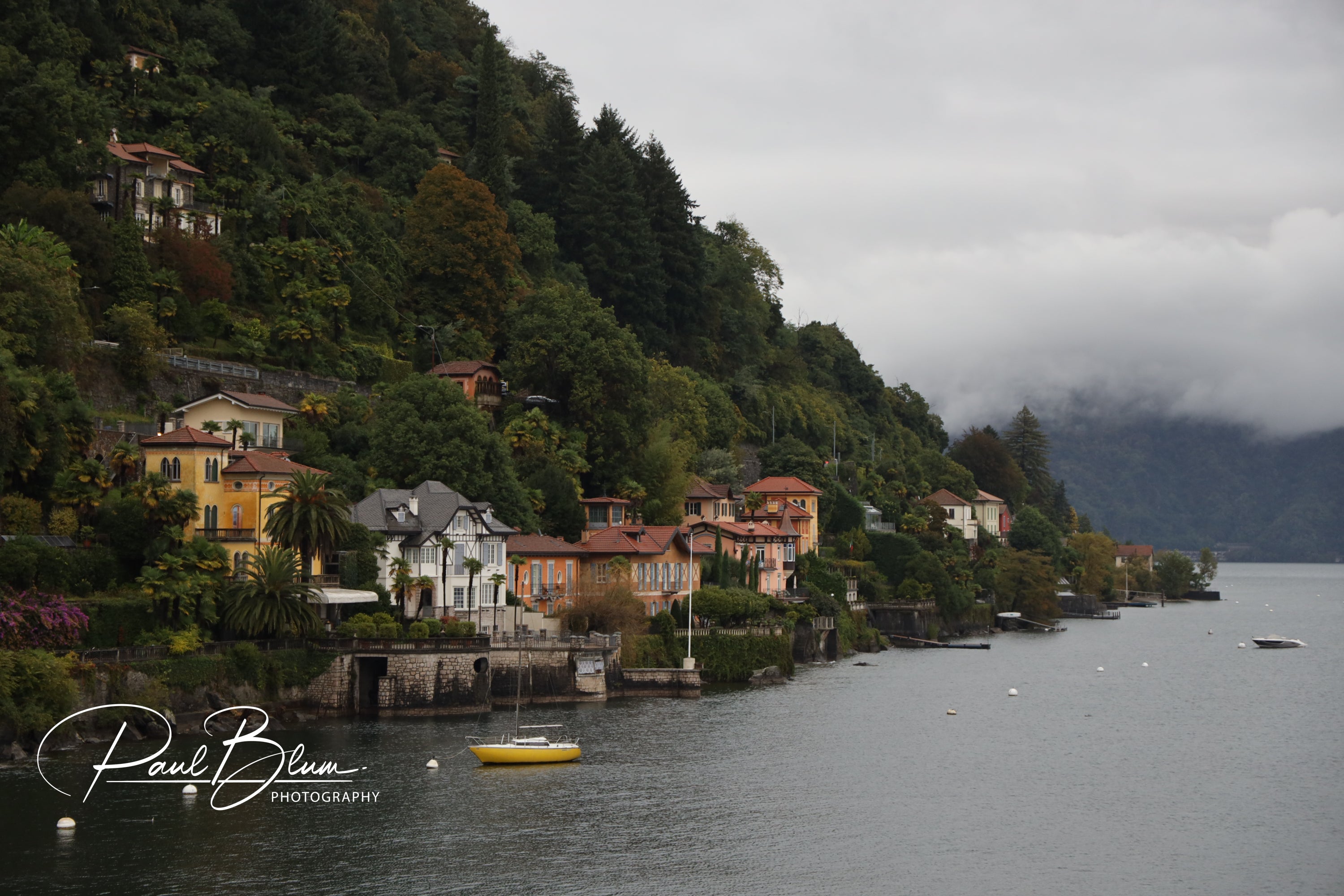 View of colourful houses along the shoreline of Lake Maggiore, Italy, with lush greenery on the hillside and a small yellow boat moored on the calm water under a cloudy sky.