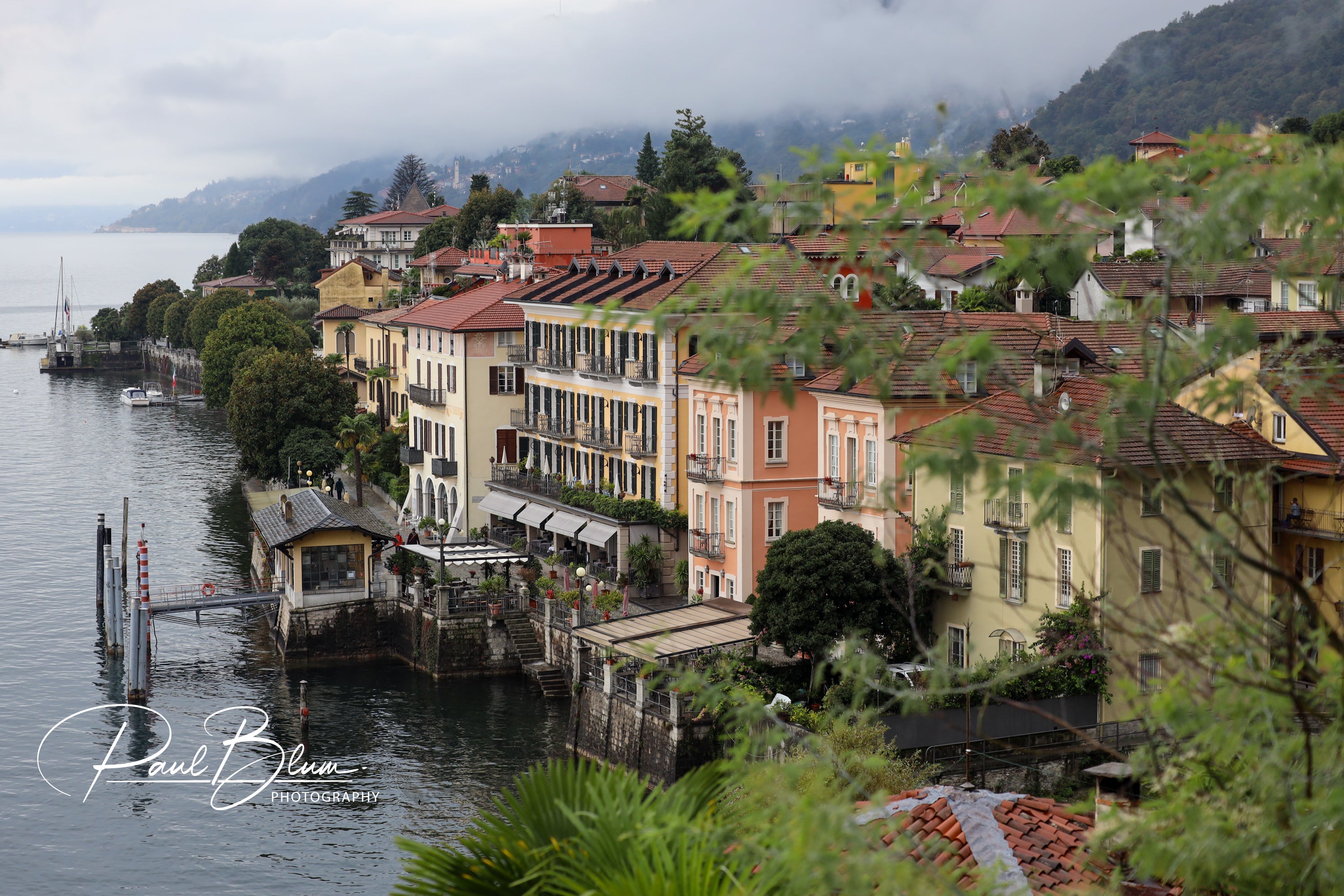 Colourful lakeside buildings in Cannero Riviera, Lake Maggiore, Italy, with a view of the waterfront and surrounding lush hills on a misty day.