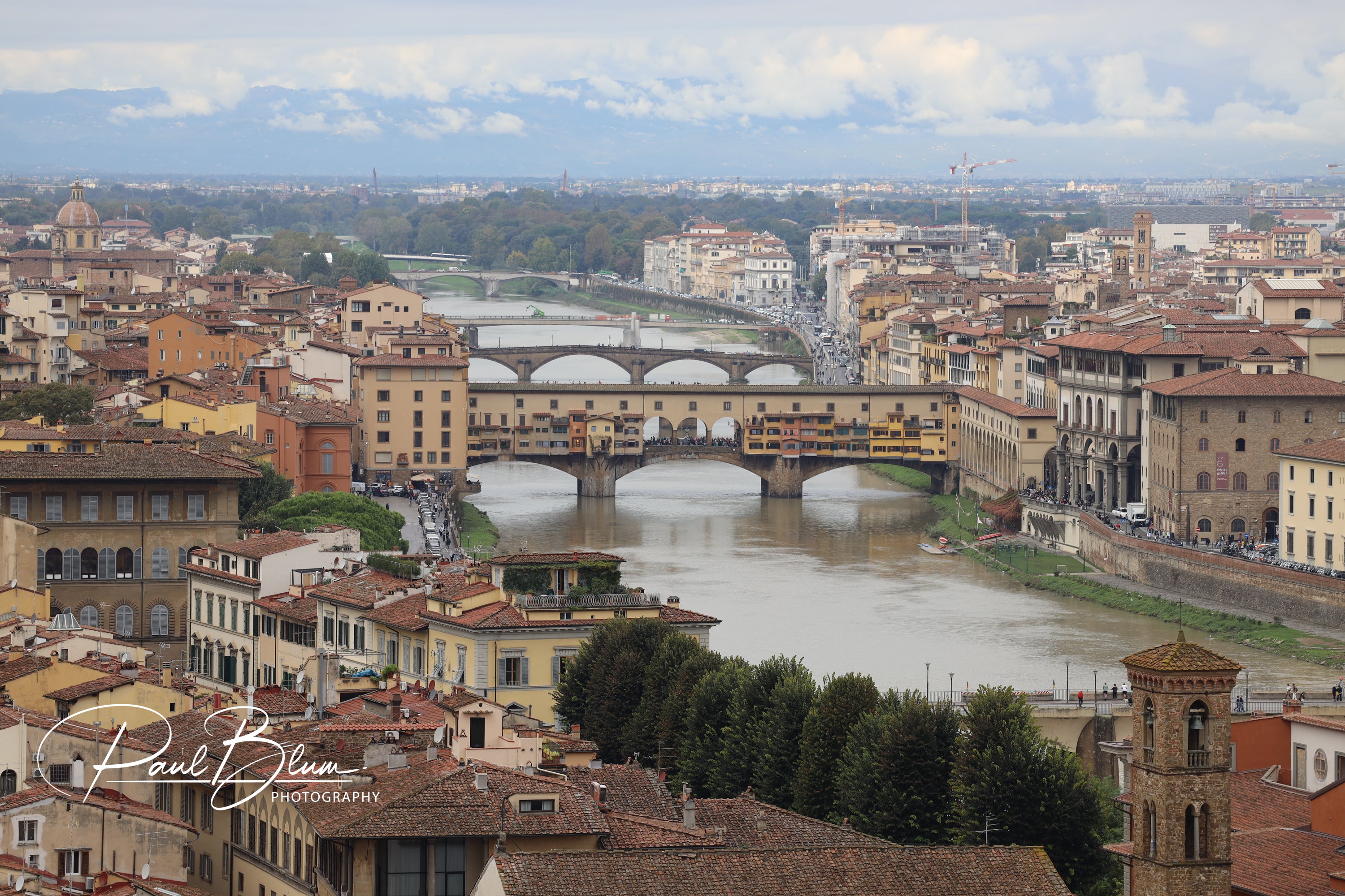 Iconic view of Florence, Italy, featuring the Ponte Vecchio bridge spanning the Arno River, surrounded by historic buildings and rooftops, with distant hills and a cloudy sky in the background.