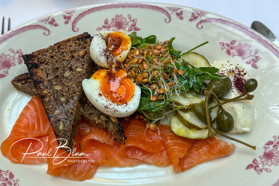 A beautifully plated breakfast featuring smoked salmon, toasted whole grain bread, soft-boiled eggs with runny yolks, sprouts, greens, pickles, capers, and a dollop of creamy cheese, captured by Paul Blum Photography.