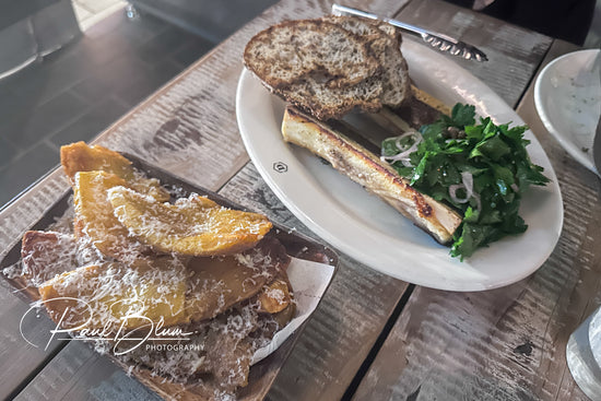 A rustic meal featuring roasted bone marrow with toasted bread and a fresh parsley salad on one plate, accompanied by a side of crispy potato wedges topped with grated cheese, captured by Paul Blum Photography.