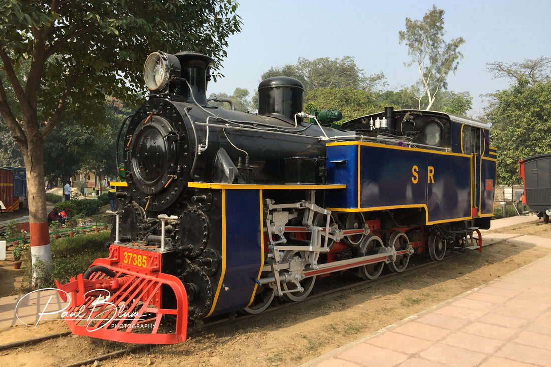 A vintage steam locomotive with a blue and black colour scheme and red detailing, captured by Paul Blum Photography. The train is displayed outdoors on a track surrounded by trees and greenery.