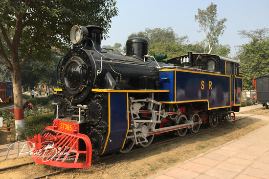 A vintage steam locomotive with a blue and black colour scheme and red detailing, captured by Paul Blum Photography. The train is displayed outdoors on a track surrounded by trees and greenery.