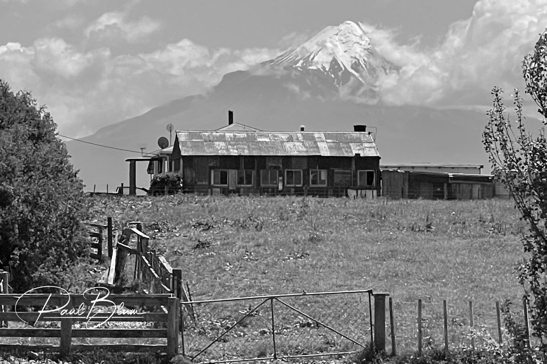 A black-and-white photograph of a rustic farmhouse with a rusted roof set against a backdrop of a snow-capped mountain peak, captured by Paul Blum Photography.