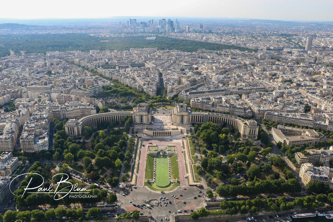 Aerial view of Trocadéro Gardens in Paris, with the Palais de Chaillot at the forefront, leading the eye towards the dense urban landscape and modern skyscrapers of La Défense in the background.