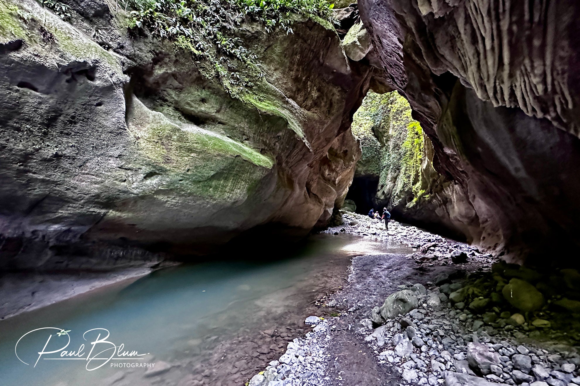 "A breathtaking view inside a large cave with rocky walls, a calm blue stream running through it, and a few people exploring the path ahead."