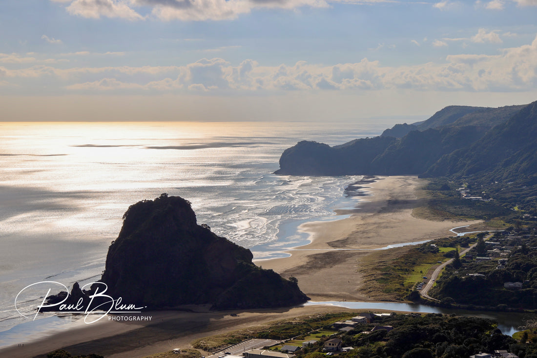 An aerial view of Piha Beach, New Zealand, showcasing a radiant sunset that casts shimmering gold reflections on the water. The image captures the dramatic black rock formation known as Lion Rock, surrounded by patterns of white surf and tidal flats. The surrounding landscape features lush green hills and a small coastal community nestled between the sea and the cliffs.