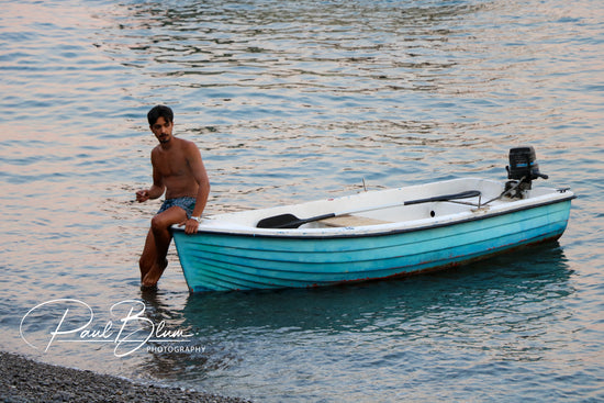 Man in swim trunks standing beside a small blue boat on the shoreline in Positano, Italy, with the evening light reflecting off the calm waters of the Amalfi Coast.