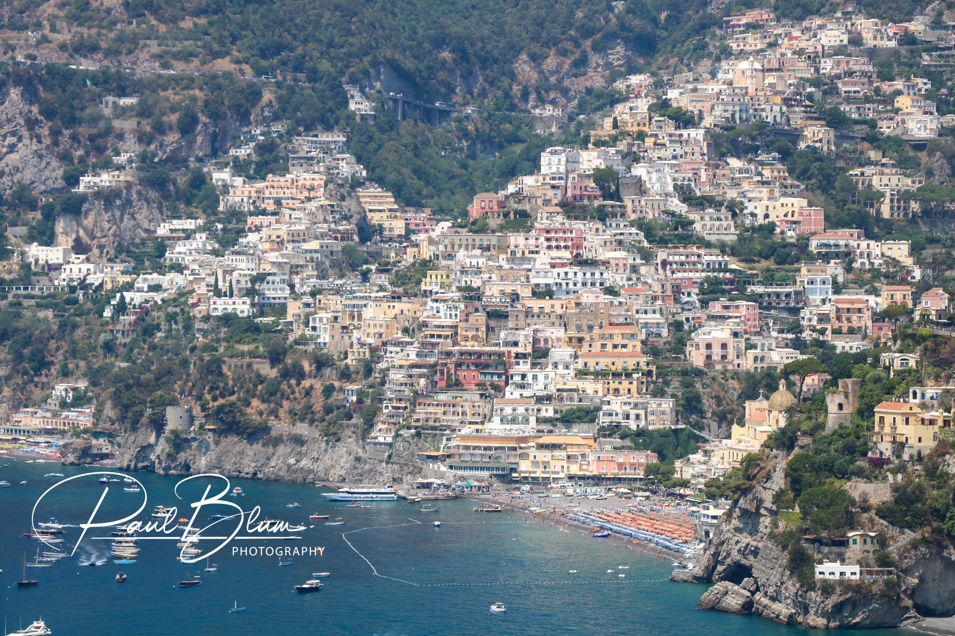 Panoramic view of Positano, Italy, with colourful buildings cascading down the steep hillside to the beach, boats moored in the Mediterranean Sea, and lush greenery on the Amalfi Coast.