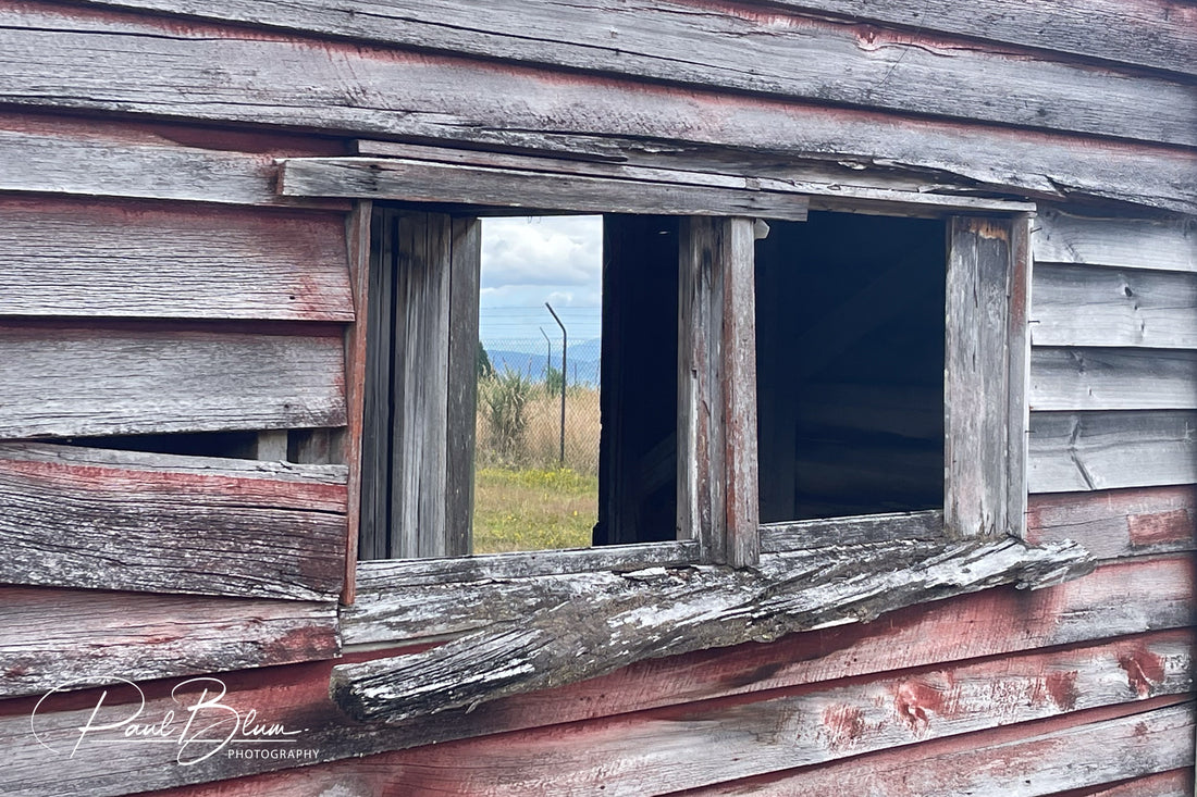 "An abandoned wooden structure with weathered and peeling paint, featuring an open window that offers a glimpse of the grassy field and distant landscape."