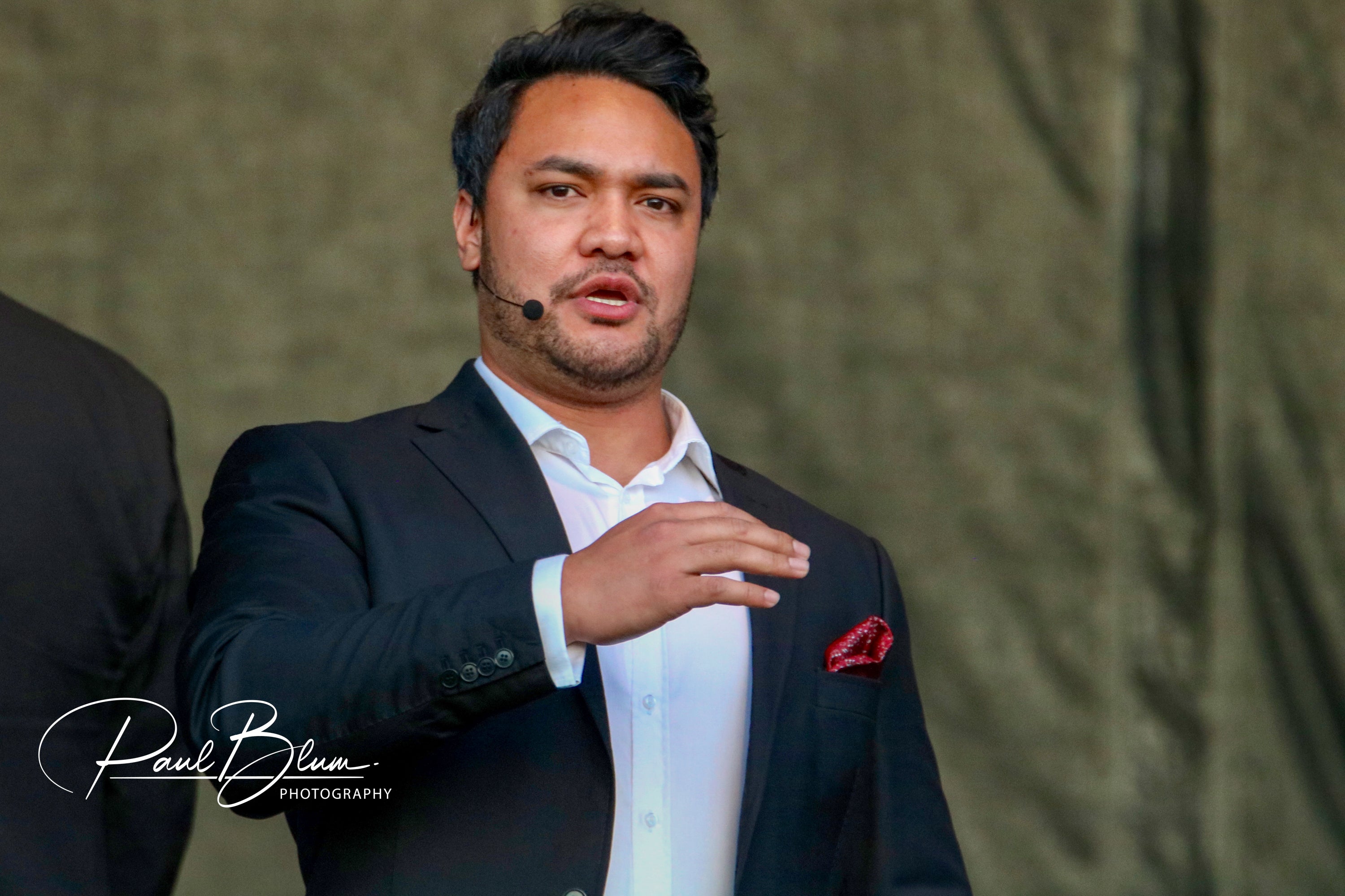 Samoan tenor Amitai Pati of Sol3 Mio engages an audience during a live performance, gesturing expressively in a dark suit, exuding charisma and vocal power.