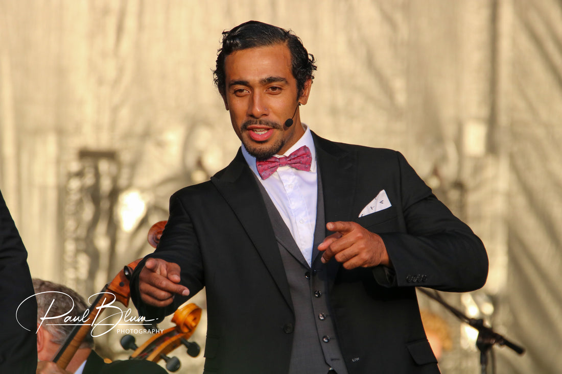 Moses Mackay, a baritone member of the New Zealand opera trio Sol3 Mio, engaging with an audience during a live outdoor performance, dressed in a formal black suit with a pink bow tie.