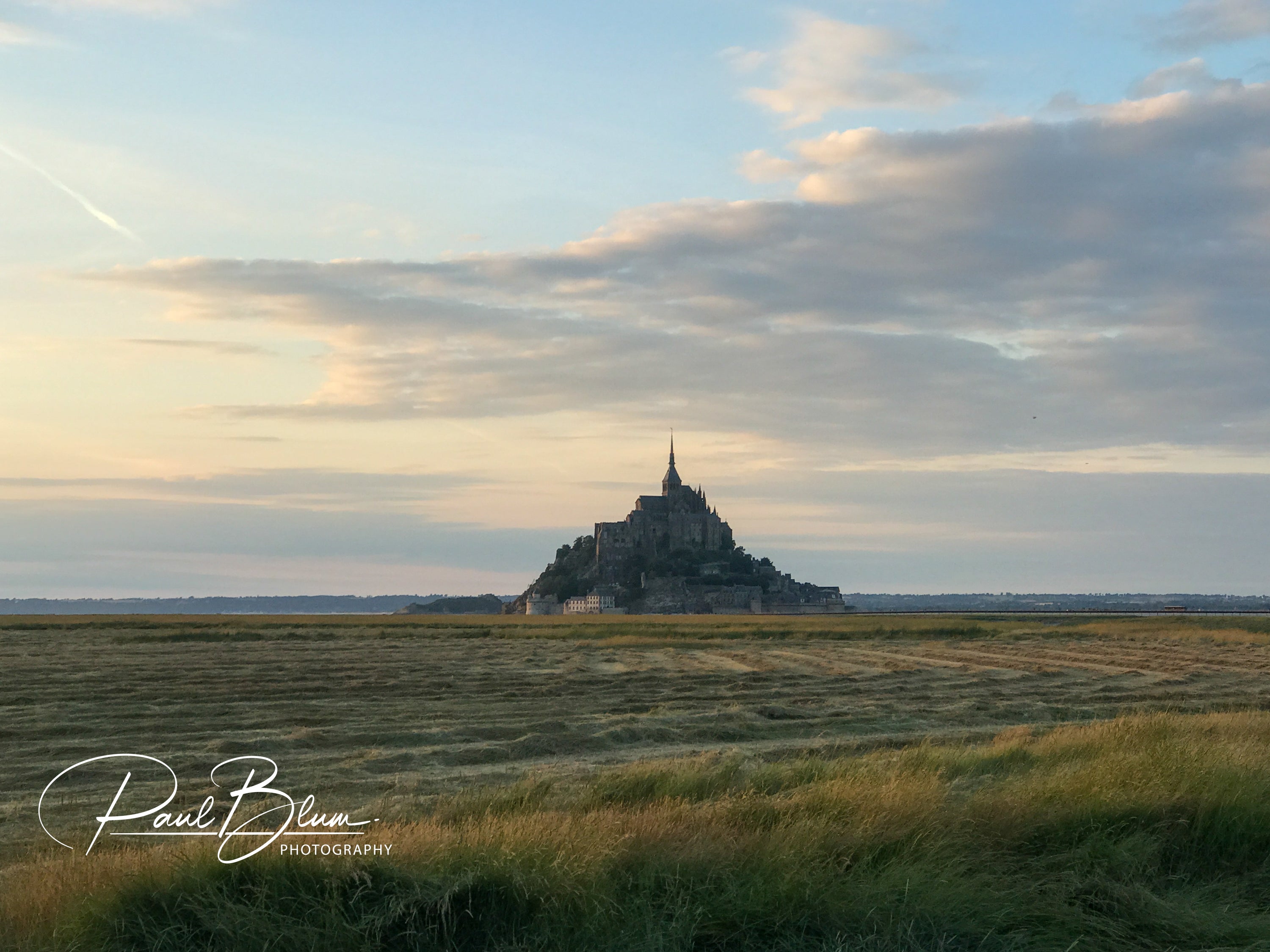 Mont Saint-Michel in France, showcasing the island’s iconic silhouette against a soft sky with fields of harvested grain in the foreground.