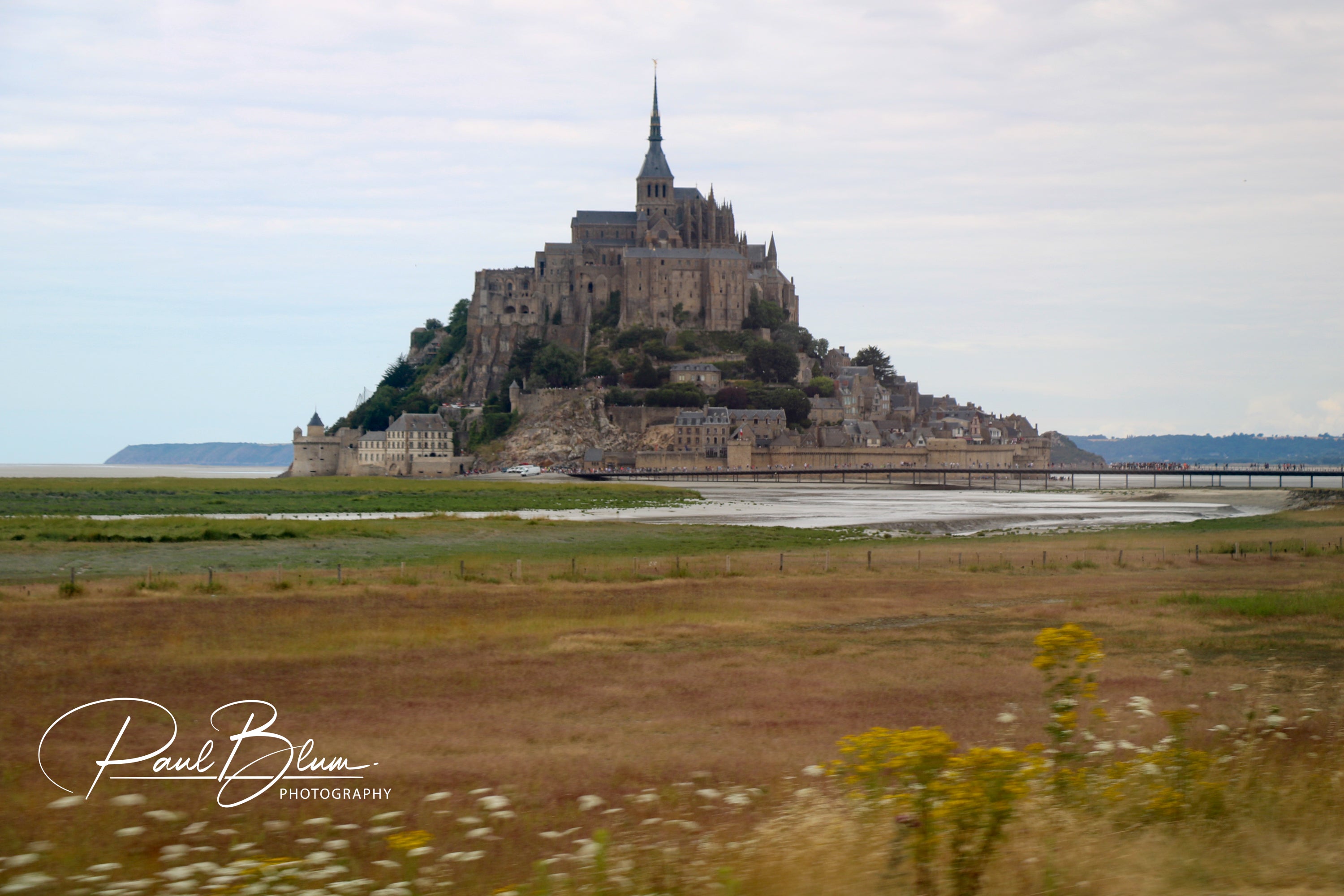 Panoramic view of Mont Saint-Michel, France, featuring the iconic island commune with its medieval abbey rising above the surrounding flat marshlands and a causeway connecting it to the mainland.