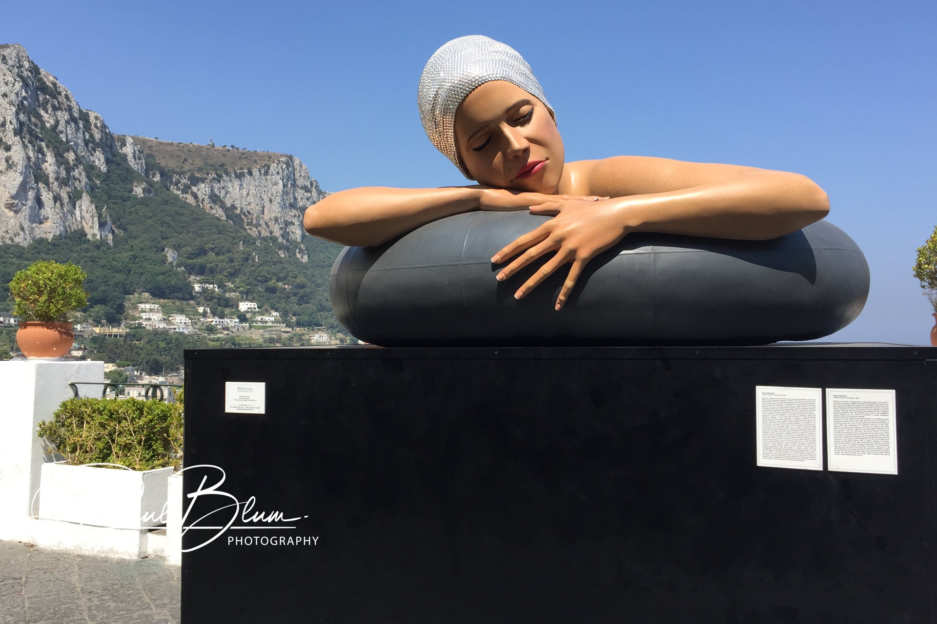 Art sculpture of a woman resting on an inflatable ring, with a serene expression, set against the backdrop of a mountain landscape in Capri, Italy.