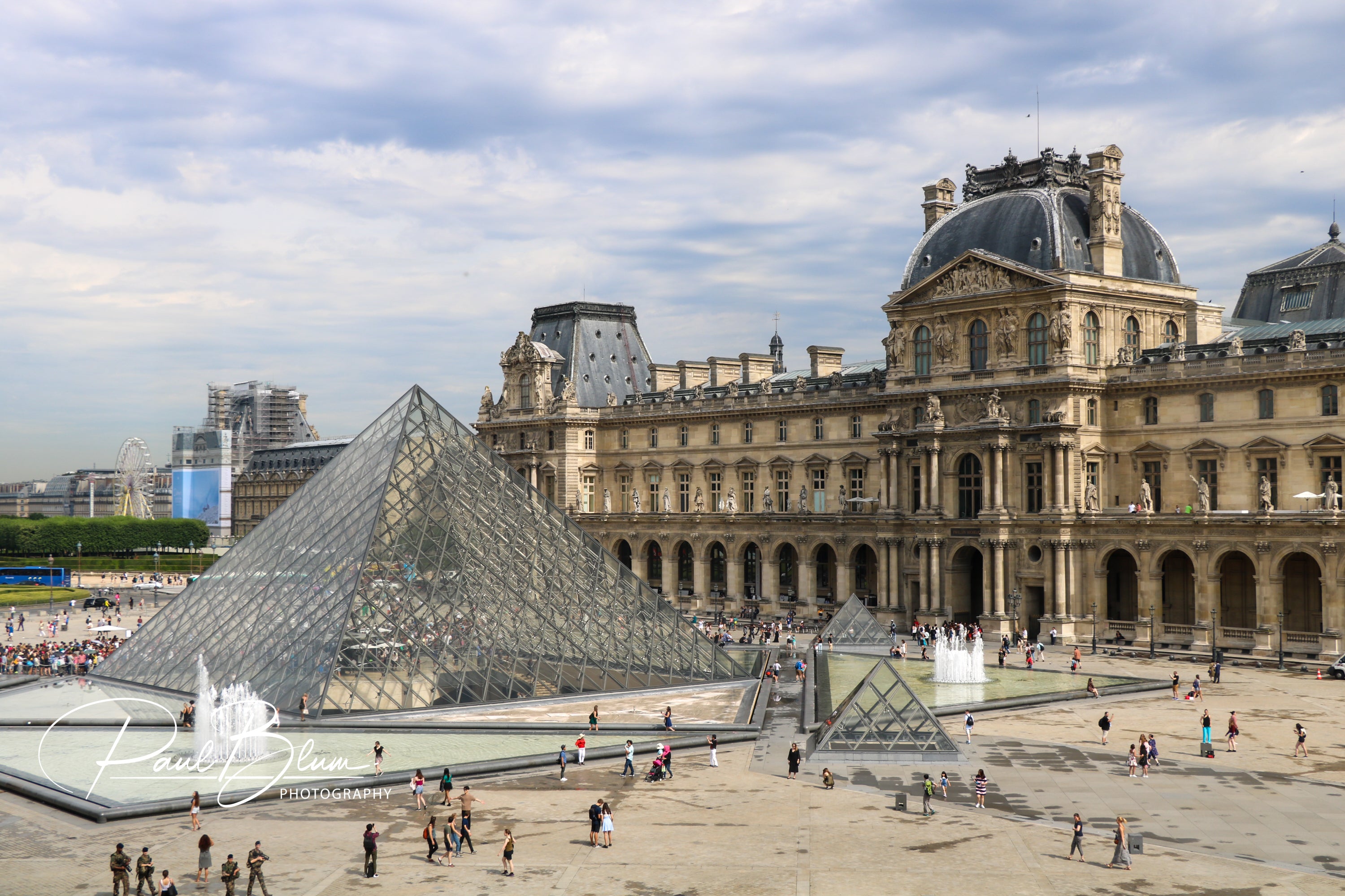 The Louvre Museum's famous glass pyramid serves as a modern centerpiece in the foreground, contrasting with the classical French Renaissance architecture of the museum buildings, under a clear sky.