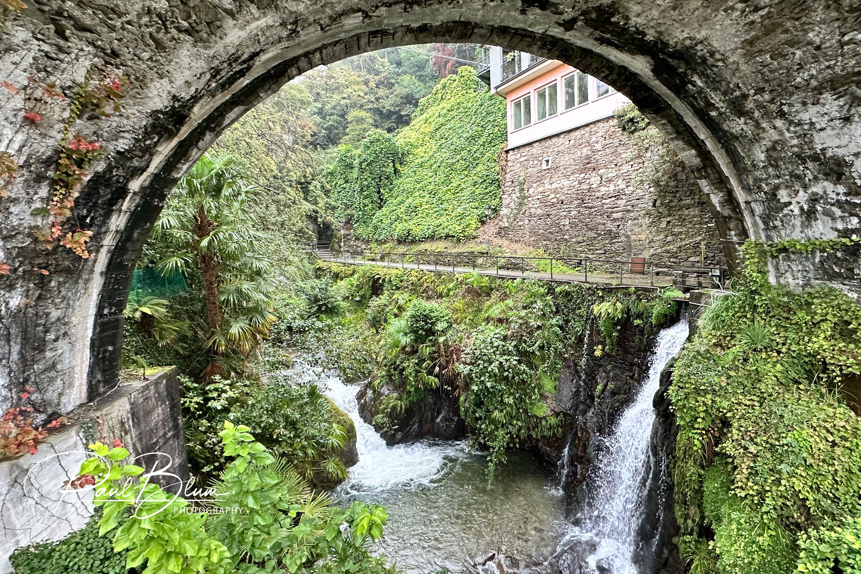 View through an old stone arch bridge in Cannero Riviera, Italy, with lush greenery and a waterfall flowing below.