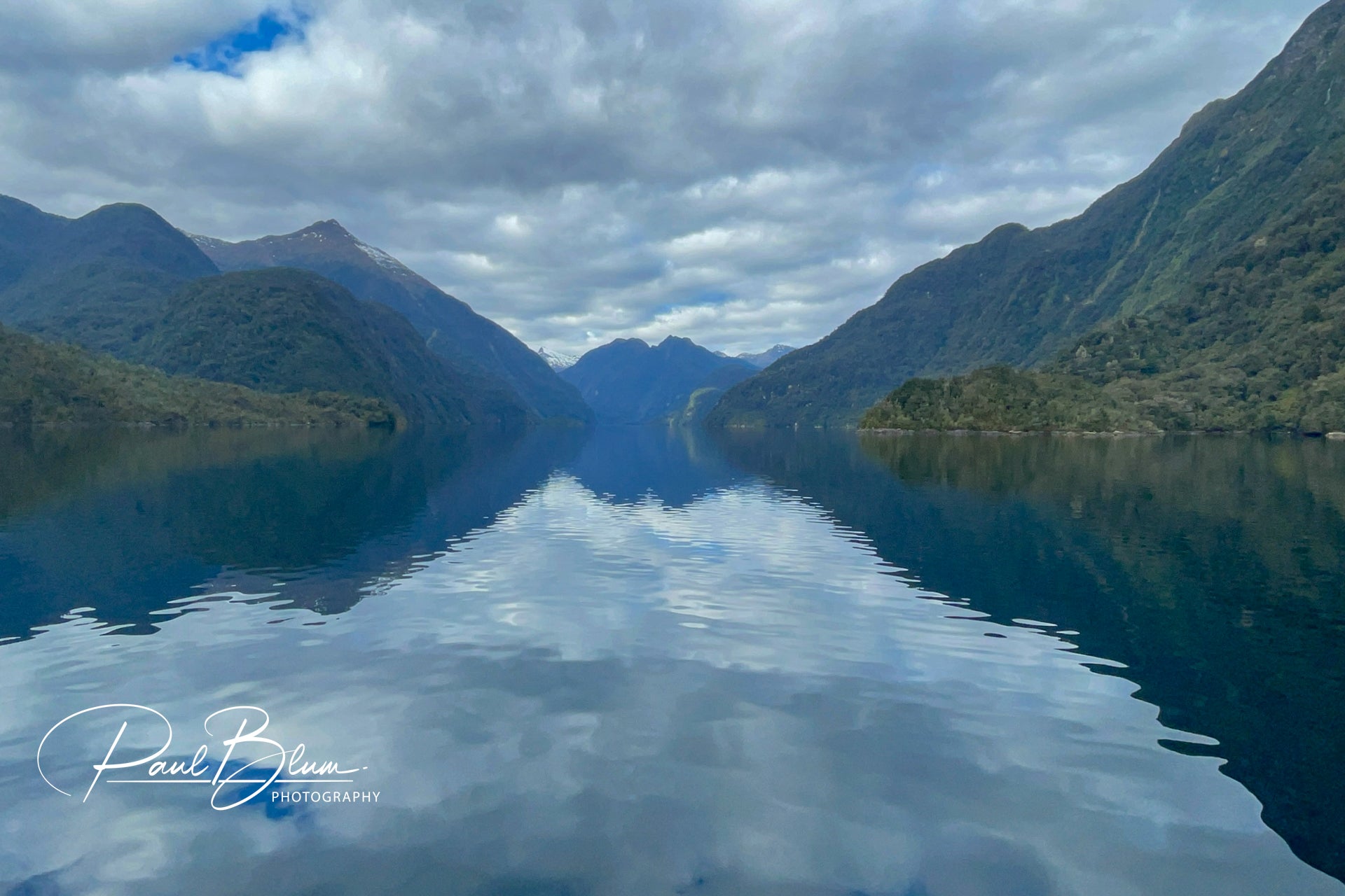 Serene view of Wet Jacket Arm in Fiordland, New Zealand, featuring calm waters flanked by lush, green mountains under a partly cloudy sky. The perfect reflections of the mountains in the still water create a symmetrical natural vista.