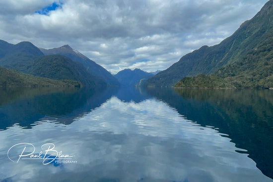 Serene view of Wet Jacket Arm in Fiordland, New Zealand, featuring calm waters flanked by lush, green mountains under a partly cloudy sky. The perfect reflections of the mountains in the still water create a symmetrical natural vista.